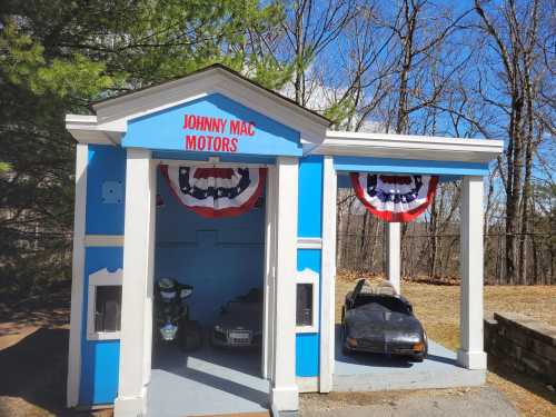 A small blue building with "Johnny Mac Motors" sign, featuring two toy cars and patriotic decorations.