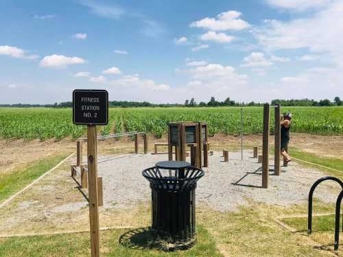 Outdoor fitness station in a field, featuring exercise equipment and a trash bin, with blue skies and crops in the background.