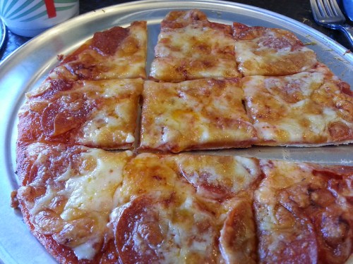 A close-up of a pizza with pepperoni and melted cheese, cut into square slices on a metal tray.