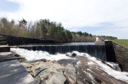 A serene waterfall cascades over a stone dam, surrounded by lush greenery and a clear blue sky.