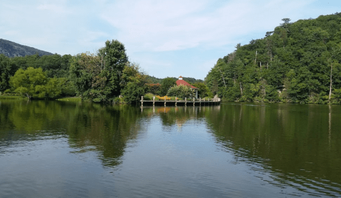 A serene lake scene with a wooden dock, surrounded by lush greenery and a distant red-roofed building.