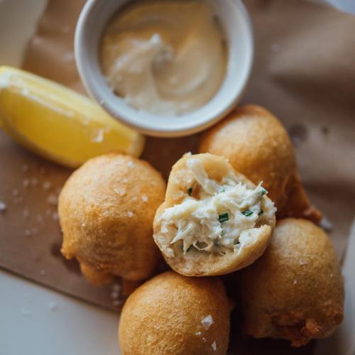 Golden fried balls filled with crab, served with a side of creamy sauce and a lemon wedge on brown paper.