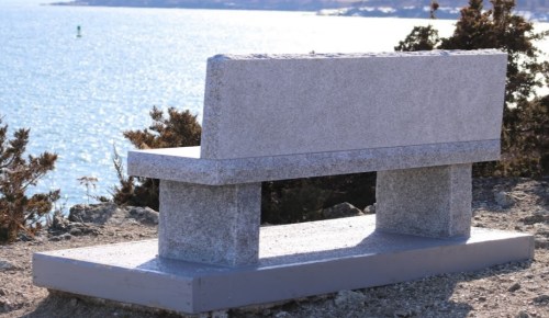 A gray stone bench overlooking a serene body of water, surrounded by sparse vegetation.