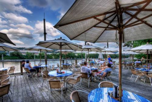 Outdoor dining area by a river, featuring tables with umbrellas and patrons enjoying the scenery.