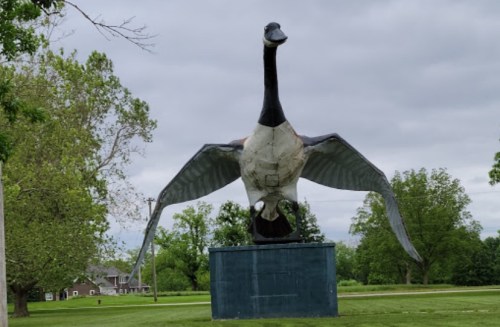 A large statue of a goose with outstretched wings, set against a cloudy sky and green trees.