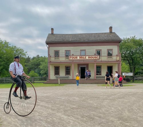 A man rides a penny-farthing bicycle in front of the historic Four Mile House, with visitors entering the building.