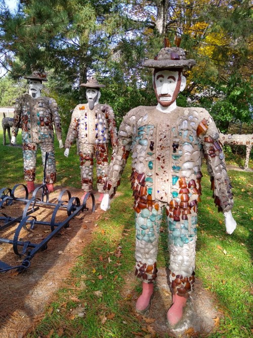 Three whimsical sculptures made of colorful glass and stone, resembling men in hats, stand in a grassy area surrounded by trees.