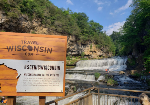 A wooden sign for Travel Wisconsin in front of a scenic waterfall and lush greenery.