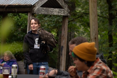 A woman holds an owl while speaking to a group seated at a table in a forested area.