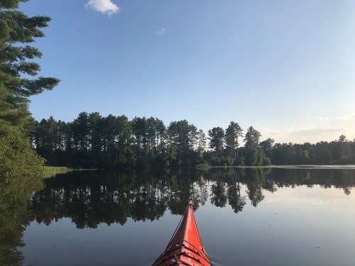 A kayak's orange bow on calm water, reflecting trees and a clear blue sky.