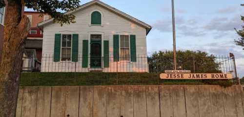 Historic white house with green shutters, labeled "Jesse James Home," set against a sunset sky.