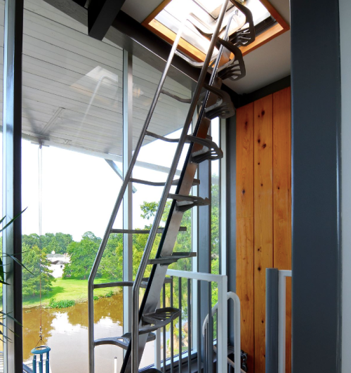 A spiral staircase leads to a skylight, with a view of greenery and water outside through large windows.