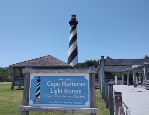 A striped lighthouse towers over a welcome sign at Cape Hatteras Light Station, surrounded by green grass and trees.