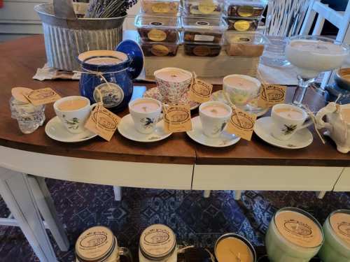 A display of decorative candles in teacups and jars, arranged on a wooden table with various labels and tags.