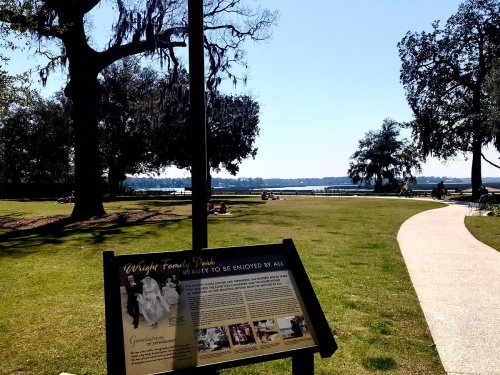 A sunny park scene with a sign about Wright Family Park, trees, and people enjoying the waterfront view.