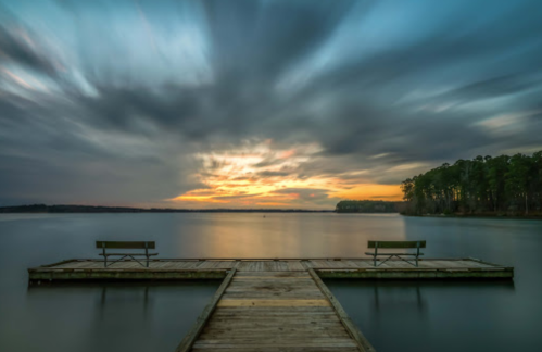 A serene lake view at sunset, featuring a wooden dock with two benches and dramatic clouds in the sky.