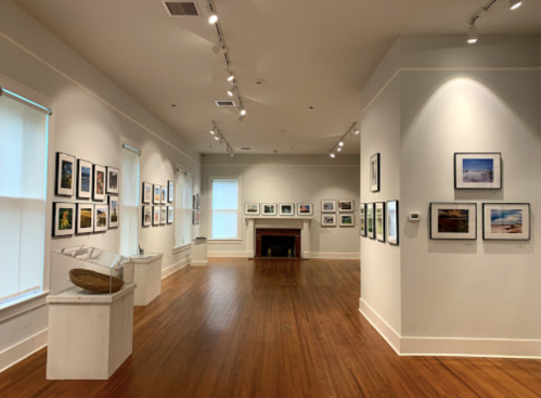 A spacious art gallery with framed photographs on the walls and a wooden floor, featuring a fireplace in the background.