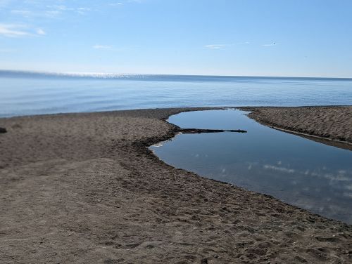 A calm beach scene with a shallow pool of water reflecting the sky, surrounded by sandy shore and distant horizon.