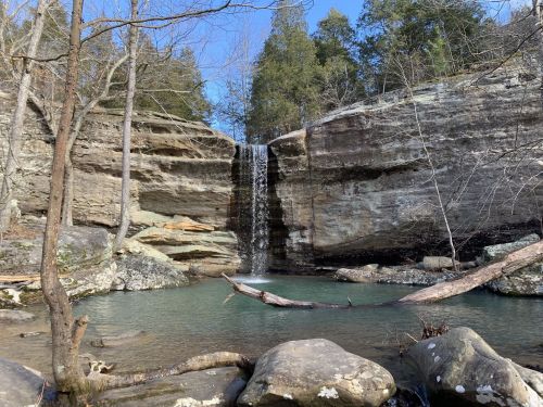 A serene waterfall cascades over rocky cliffs into a clear pool, surrounded by trees and boulders under a blue sky.