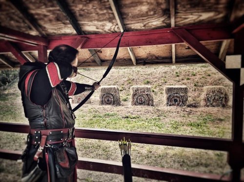 A person in medieval attire aims a bow at targets made of hay bales in an outdoor archery range.