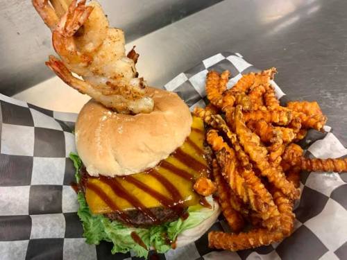 A burger with cheese, lettuce, and shrimp on top, served with curly fries in a checkered basket.