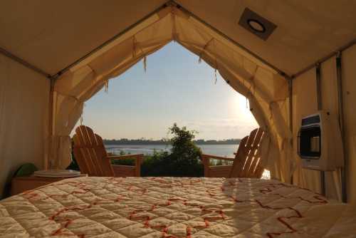 View from inside a tent, showcasing a cozy bed and two chairs overlooking a serene lake at sunrise.