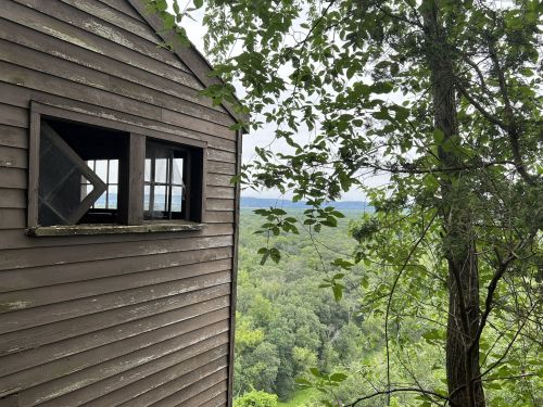 A weathered wooden building with a broken window overlooks a lush green landscape and distant hills.