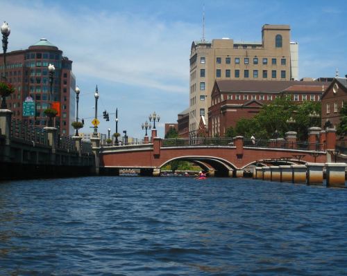 A view of a river with a brick bridge and buildings lining the banks under a clear blue sky.