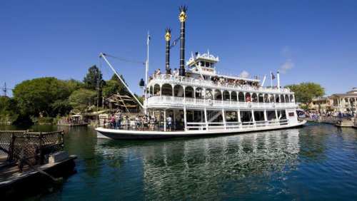 A large steamboat with multiple decks sails on a calm river under a clear blue sky, surrounded by greenery.