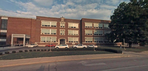 A large brick school building with multiple windows, parked cars in front, and a cloudy sky above.