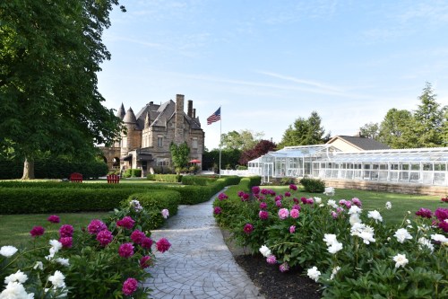 A historic mansion with an American flag, surrounded by blooming flowers and greenhouses in a landscaped garden.