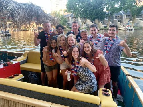 A group of friends poses on a boat, wearing leis and smiling, with a scenic waterfront in the background.