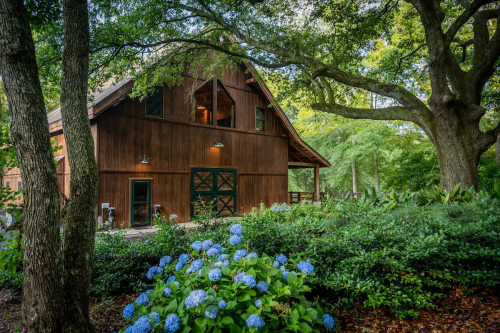 A rustic wooden barn surrounded by lush greenery and blue hydrangeas under a clear sky.