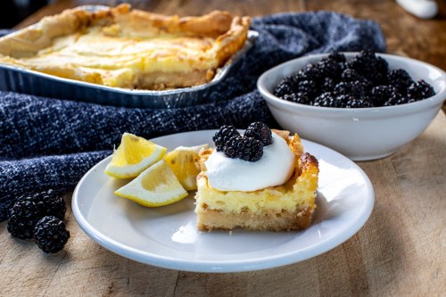 A slice of creamy dessert topped with blackberries and whipped cream, served with lemon wedges, beside a bowl of blackberries.