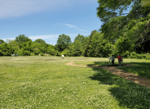 A grassy field with trees in the background, featuring two people near a path and a white circular object in the distance.