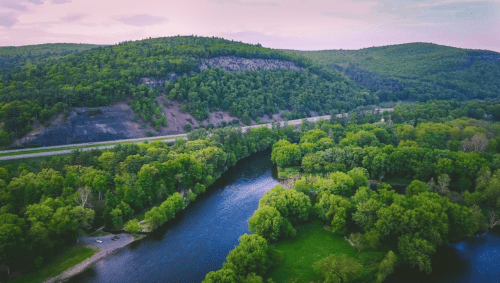 Aerial view of a winding river surrounded by lush green trees and rolling hills under a soft pink sky.