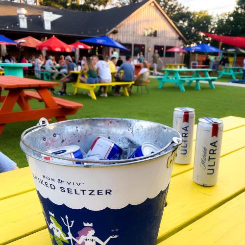 A bucket of spiked seltzer sits on a bright yellow table, with people enjoying drinks in a lively outdoor setting.