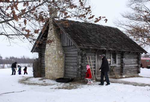 A snowy scene featuring a log cabin with a stone chimney, where a family is playing outside.
