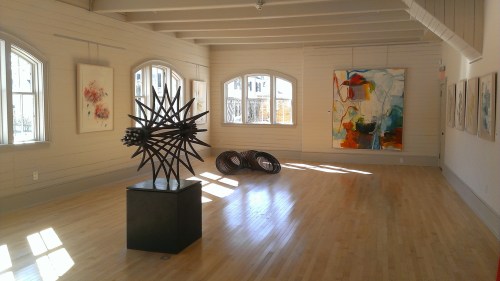 Art gallery interior featuring abstract sculptures and colorful paintings, with natural light streaming through windows.