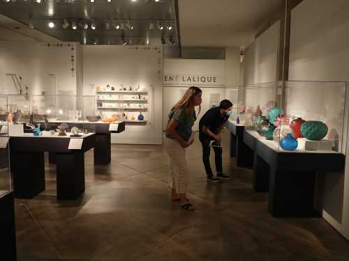 Two visitors examine colorful glass sculptures in a museum gallery featuring Lalique's work.