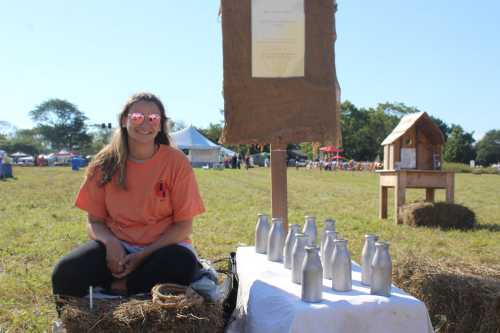 A smiling person in sunglasses sits on hay, with milk bottles displayed on a table at an outdoor event.