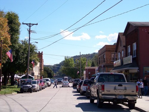 A small town street lined with parked trucks, shops, and trees under a clear blue sky with a few clouds.