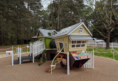 Colorful playground structure resembling two houses, with slides, climbing features, and a fenced area in a wooded setting.