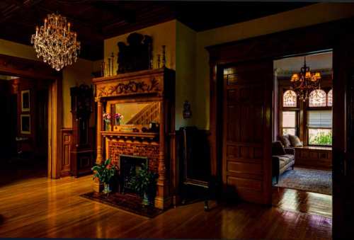 A dimly lit room featuring a decorative fireplace, wooden accents, and a view of a cozy sitting area through an open door.