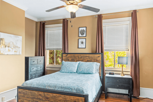 Cozy bedroom with a wooden bed, blue bedding, nightstand, and large windows with brown curtains.