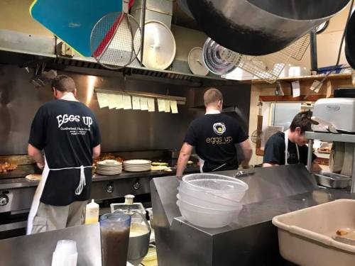 Three cooks work in a busy kitchen, preparing food at a grill with various utensils and plates around them.