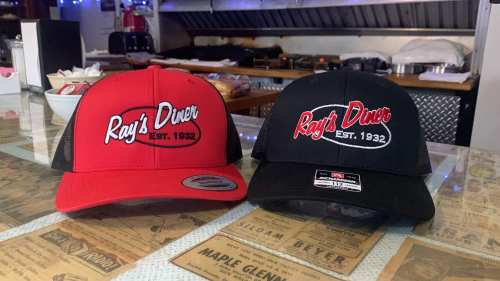 Two baseball caps on a table: one red with "Ray's Diner EST. 1932" and one black with the same logo.