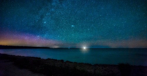 A serene night sky filled with stars over a calm ocean, with a faint glow on the horizon and a sandy shoreline.