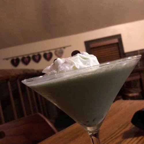 A close-up of a green cocktail topped with whipped cream and a cherry, set on a wooden table in a cozy setting.