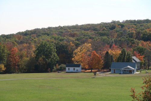 A scenic view of a green field with two houses, surrounded by vibrant autumn trees on a hillside.
